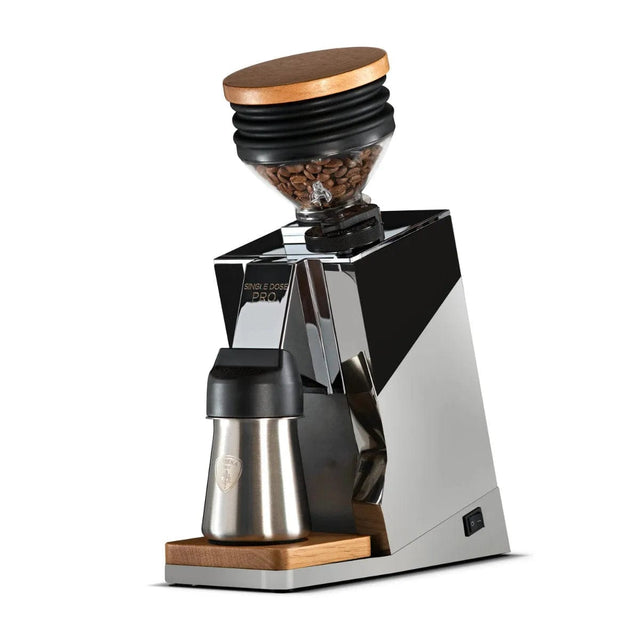 The Eureka Oro Mignon Single Dose Pro Grinder in chrome is a sleek coffee grinder featuring flat burrs and a transparent bean hopper topped with a wooden lid. It boasts a zero-retention design and includes a stainless steel cup for grounds collection, all elegantly set on a wooden platform. The modern and minimalist appearance makes it truly striking.