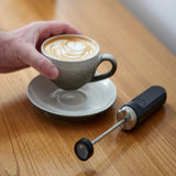 A hand holds a gray coffee cup with latte art on a saucer atop a wooden table. Next to it rests the Subminimal V2 NanoFoamer (Black) by Subminimal, ideal for creating smooth microfoam at home.