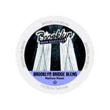 A circular label for Brooklyn Bean's medium roast coffee pods. The text reads, "Brooklyn Bean" at the top and "Brooklyn Bridge Blend Medium Roast Single-Serve Pods" at the bottom over a black-and-white image of the Brooklyn Bridge. "BROOKLYN BEAN" is repeated around the edge. Compatible with Keurig brewers.