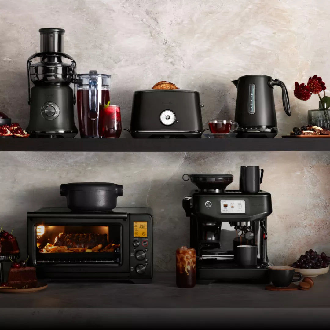 Two kitchen shelves showcase a juicer, blender, toaster, and the Breville the Smart Kettle Luxe 1.7L BKE845BTR (Black Truffle) on top. Below are a toaster oven and espresso machine, surrounded by drinks, bread, and desserts.