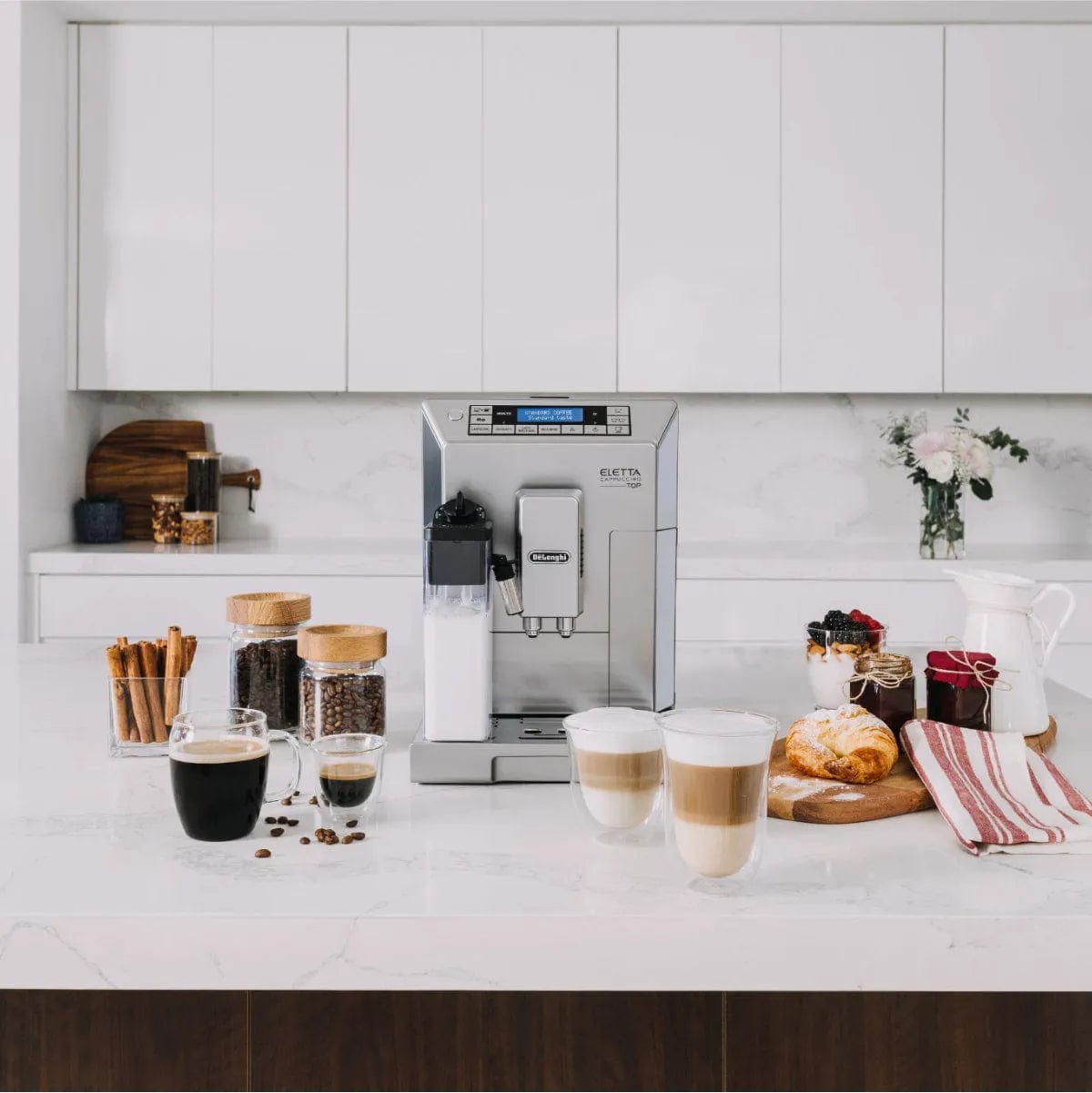 A refurbished DeLonghi Eletta Cappuccino Top Super Automatic Espresso Machine ECAM45760S (Silver) sits on a clean, organized white kitchen counter with coffee beans, cups, pastries, a milk jug, and flowers in the background.