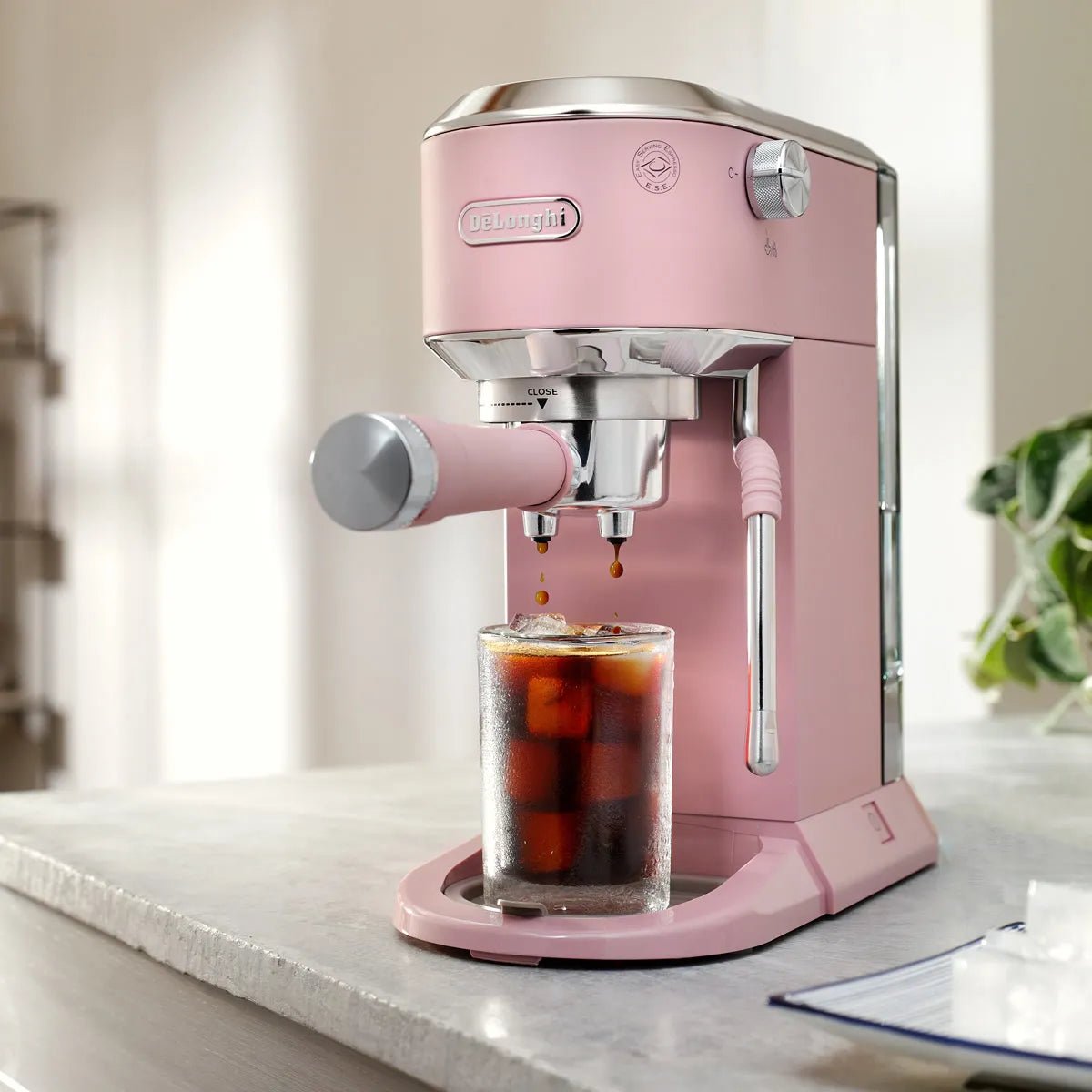 A DeLonghi Dedica Duo Espresso Machine with Cold Brew EC890PK (Rosé) brews iced coffee into a glass of ice cubes on a kitchen counter, beside a plant and in front of a blurred background.