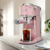 A DeLonghi Dedica Duo Espresso Machine with Cold Brew EC890PK (Rosé) brews iced coffee into a glass of ice cubes on a kitchen counter, beside a plant and in front of a blurred background.