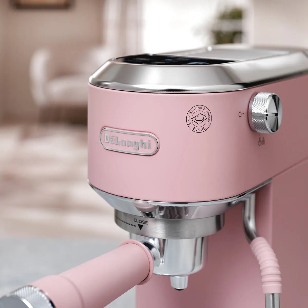 Close-up of the DeLonghi Dedica Duo Espresso Machine with Cold Brew EC890PK (Rosé) on backorder, showing its control knob, logo, and portafilter handle in a softly lit kitchen.