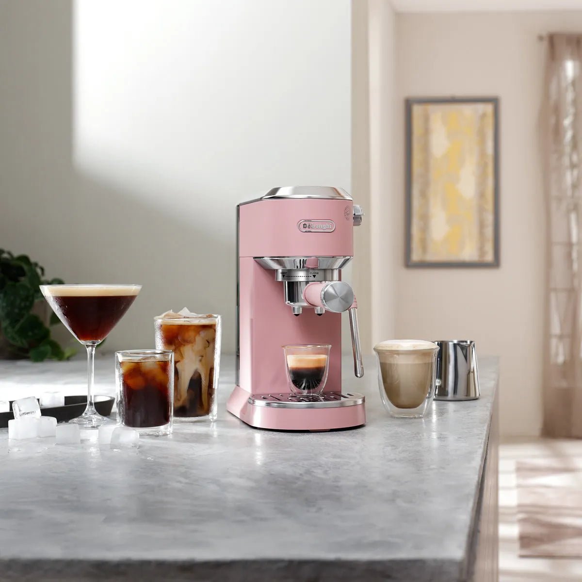 A DeLonghi Dedica Duo Espresso Machine with Cold Brew EC890PK (Rosé) sits on a kitchen counter, surrounded by four coffee drinks and a metal milk frothing pitcher, as sunlight streams through a window in the background.
