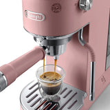 The DeLonghi Dedica Duo Espresso Machine with Cold Brew EC890PK (Rosé) brews two streams of coffee into a clear glass cup on its metallic drip tray.