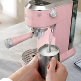 A person froths milk in a stainless steel pitcher using the DeLonghi Dedica Duo Espresso Machine with Cold Brew EC890PK (Rosé) on a light countertop.