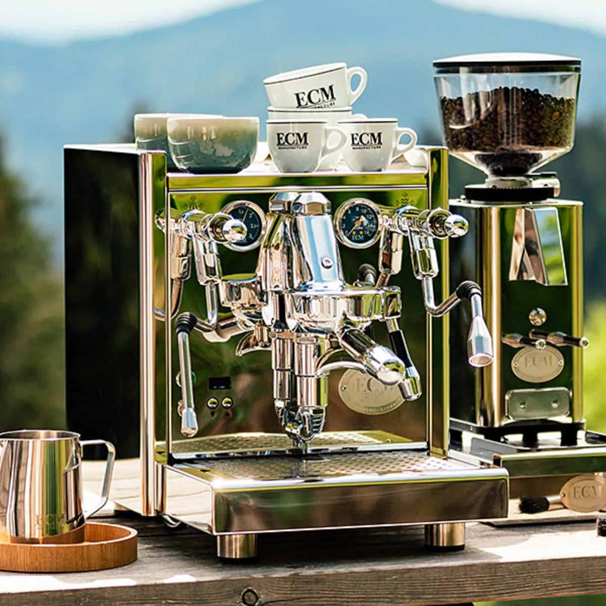 An ECM Technika VI Dual Boiler Espresso Machine with PID from ECM, topped with mugs, sits on a wooden outdoor surface beside a matching ECM grinder filled with beans, all framed by trees and mountains in the background.