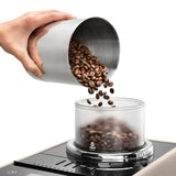 A hand pours coffee beans from a metal container into the bean hopper of the De’Longhi Rivelia Super Automatic Espresso Machine EXAM44055BG (Sand Beige) by DeLonghi, featuring the innovative Bean Switch System for easy flavor changes.