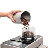 A hand pours coffee beans from a metal cup into the hopper of the De’Longhi Rivelia Super Automatic Espresso Machine EXAM44055BG (Sand Beige) by DeLonghi, featuring a Bean Switch System, set against a white background.