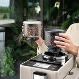 A person holds two containers filled with coffee beans, placing one onto the DeLonghi Rivelia Super Automatic Espresso Machine EXAM44055BG (Sand Beige), which features a Bean Switch System, in a bright, plant-filled room.
