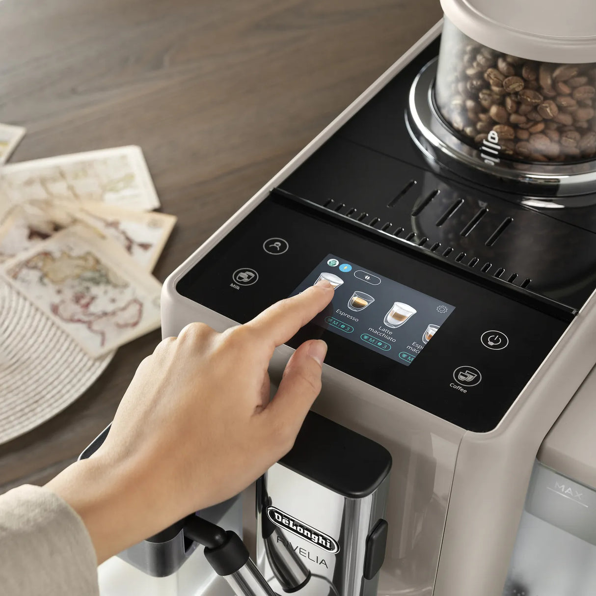 A hand selects a coffee option on the touchscreen display of the De’Longhi Rivelia Super Automatic Espresso Machine EXAM44055BG in Sand Beige, featuring LatteCrema Hot Technology. Nearby, DeLonghi coffee beans are visible in the grinder alongside scattered sheets of paper atop a wooden surface.
