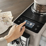 A hand selects a coffee option on the touchscreen display of the De’Longhi Rivelia Super Automatic Espresso Machine EXAM44055BG in Sand Beige, featuring LatteCrema Hot Technology. Nearby, DeLonghi coffee beans are visible in the grinder alongside scattered sheets of paper atop a wooden surface.