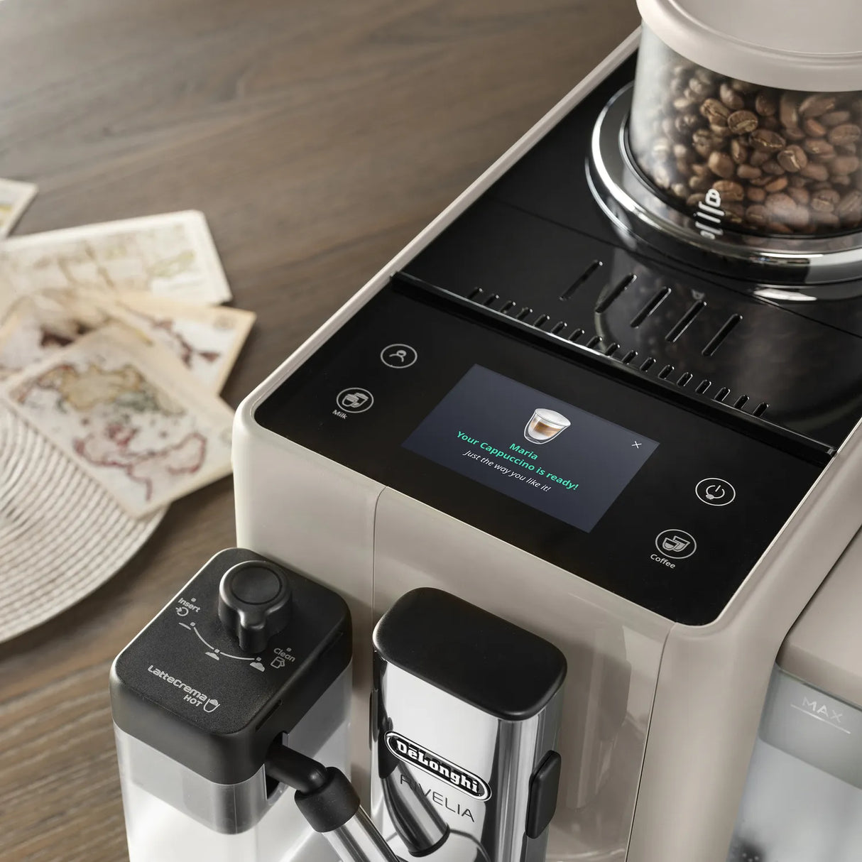 A close-up of the DeLonghi Rivelia Super Automatic Espresso Machine EXAM44055BG in Sand Beige showcases coffee beans inside, highlighting its innovative Bean Switch System and a touch screen displaying “Making a macchiato,” set on a wooden surface beside scattered papers and envelopes.