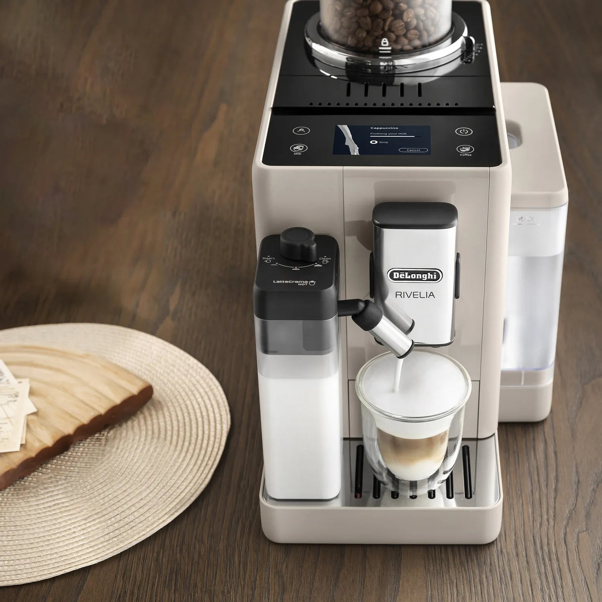 The De’Longhi Rivelia Super Automatic Espresso Machine EXAM44055BG (Sand Beige) dispenses frothed milk—using its advanced LatteCrema Hot Technology—into a glass mug, with coffee beans visible in the top compartment thanks to DeLonghi’s innovative Bean Switch System. Sliced bread rests on a woven placemat beside the machine on a wooden table.