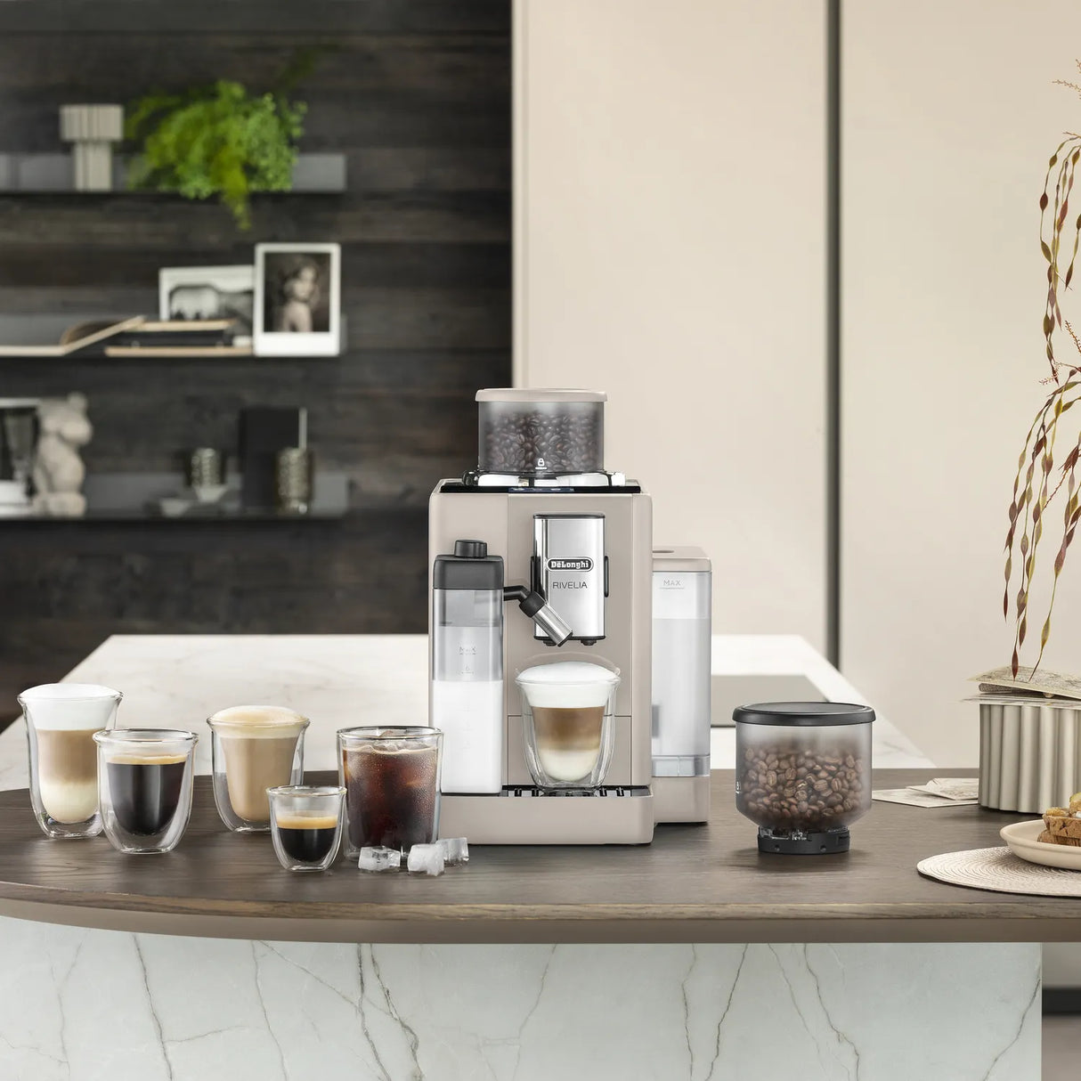 A De’Longhi Rivelia Super Automatic Espresso Machine EXAM44055BG (Sand Beige) by DeLonghi sits on a kitchen counter, surrounded by various coffee drinks in glass cups, a container of coffee beans highlighting the innovative Bean Switch System, an ice tray, and kitchen decor in the background.