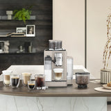 A De’Longhi Rivelia Super Automatic Espresso Machine EXAM44055BG (Sand Beige) by DeLonghi sits on a kitchen counter, surrounded by various coffee drinks in glass cups, a container of coffee beans highlighting the innovative Bean Switch System, an ice tray, and kitchen decor in the background.