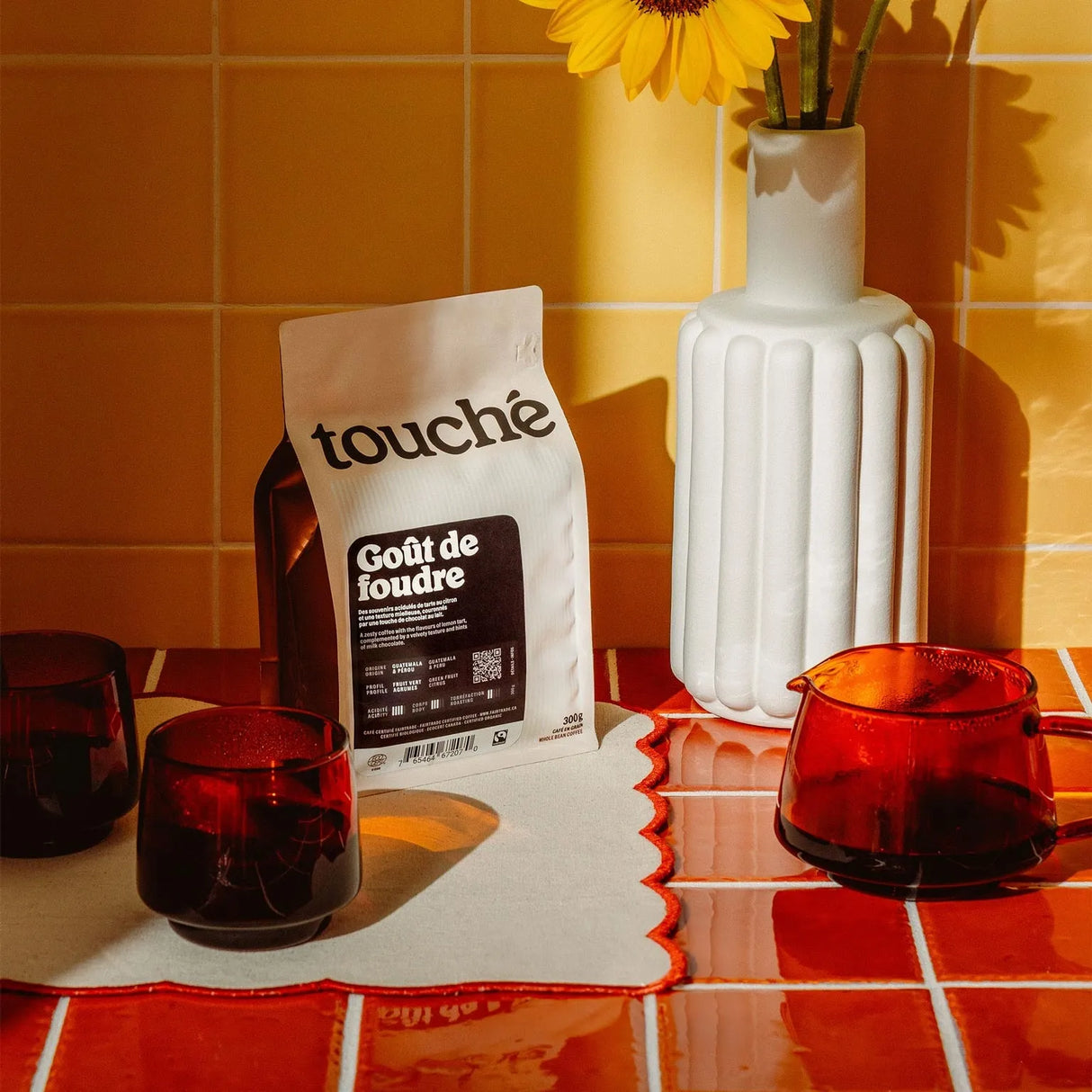 A 1 KG bag of Cafe Touche Gout de Foudre Whole Bean Coffee sits on a tiled counter alongside red glasses, a matching pitcher, a white vase with a sunflower, and yellow tiled walls in the background.