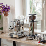 An ECM Mechanika Max Espresso Machine with Eureka Zero Grinder (Black w/ Black Spout) Bundle by ECM sits by a window with cups and purple flowers, creating a cozy spot for espresso with precise brew temperature control.