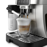 The De'Longhi Magnifica Start Super Automatic Espresso Machine with LatteCrema System ECAM22080SB (Silver) expertly pours coffee into a glass cup, creating a beautifully layered cappuccino. Equipped with DeLonghi’s innovative LatteCrema System, it delivers perfectly textured milk every time. A milk frother is attached on the left side, dispensing creamy foam into a measuring container. The espresso machine is positioned on a sleek metal drip tray.