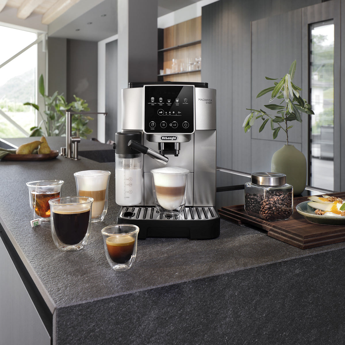 A DeLonghi Magnifica Start Super Automatic Espresso Machine with LatteCrema System ECAM22080SB (Silver) sits on a kitchen counter, accompanied by five cups showcasing different coffee drinks, a jar of coffee beans, and a potted plant. The setup’s modern aesthetic is elevated by the machine’s integrated conical burr grinder from DeLonghi, ensuring freshly ground beans for the perfect brew every time.