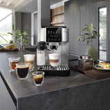 A DeLonghi Magnifica Start Super Automatic Espresso Machine with LatteCrema System ECAM22080SB (Silver) sits on a kitchen counter, accompanied by five cups showcasing different coffee drinks, a jar of coffee beans, and a potted plant. The setup’s modern aesthetic is elevated by the machine’s integrated conical burr grinder from DeLonghi, ensuring freshly ground beans for the perfect brew every time.