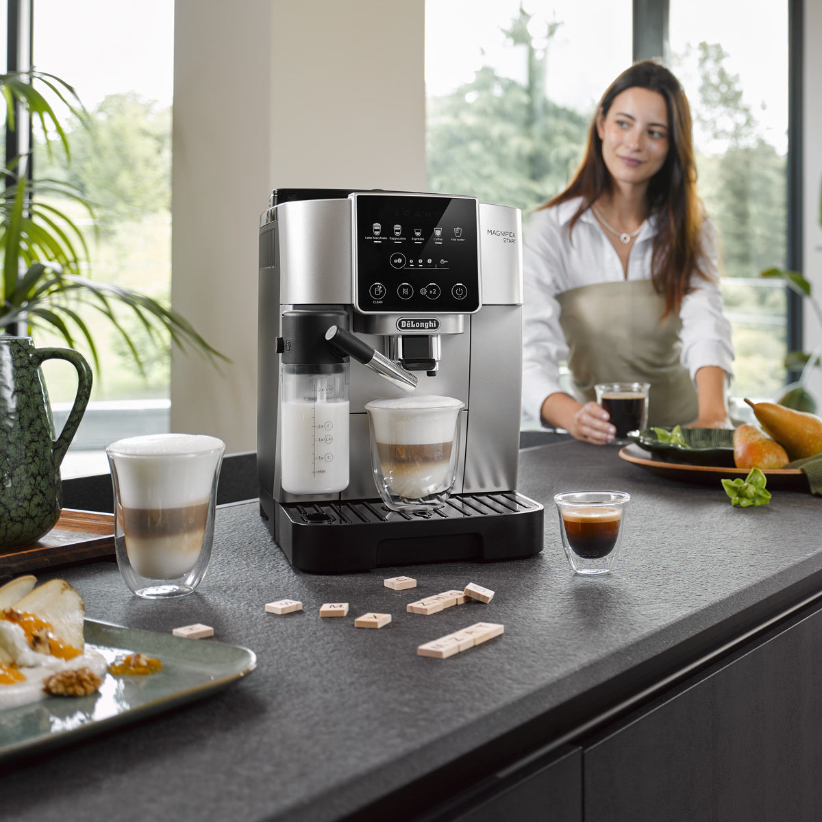 A woman stands by a kitchen counter with a DeLonghi Magnifica Start Super Automatic Espresso Machine with LatteCrema System ECAM22080SB in silver, expertly preparing coffee drinks. Glasses of coffee, pastries, and fruit are arranged on the counter near her in a bright, modern kitchen.