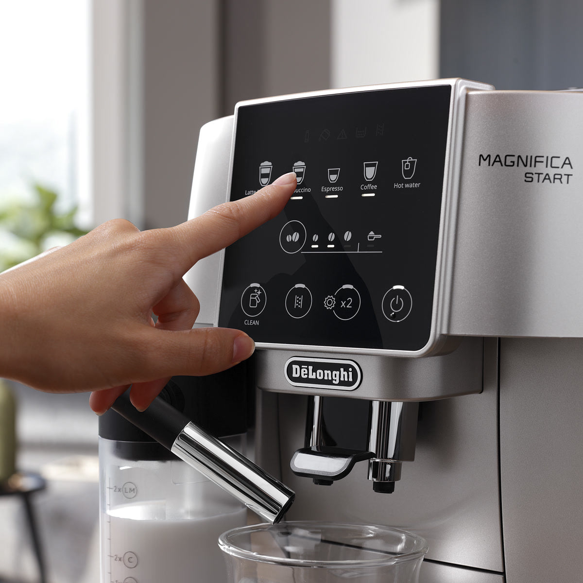 A hand presses the "Espresso" button on a DeLonghi Magnifica Start Super Automatic Espresso Machine with LatteCrema System ECAM22080SB (Silver), featuring a digital display. A glass sits beneath the coffee spout, and the LatteCrema milk frother is visible on the left.