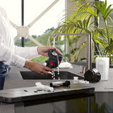 A person rinses a part of the DeLonghi Magnifica Start Super Automatic Espresso Machine with LatteCrema System ECAM22080SB (Silver) under a modern kitchen faucet, with various disassembled components and tools on the countertop beside them. Green plants and large windows are visible in the background.