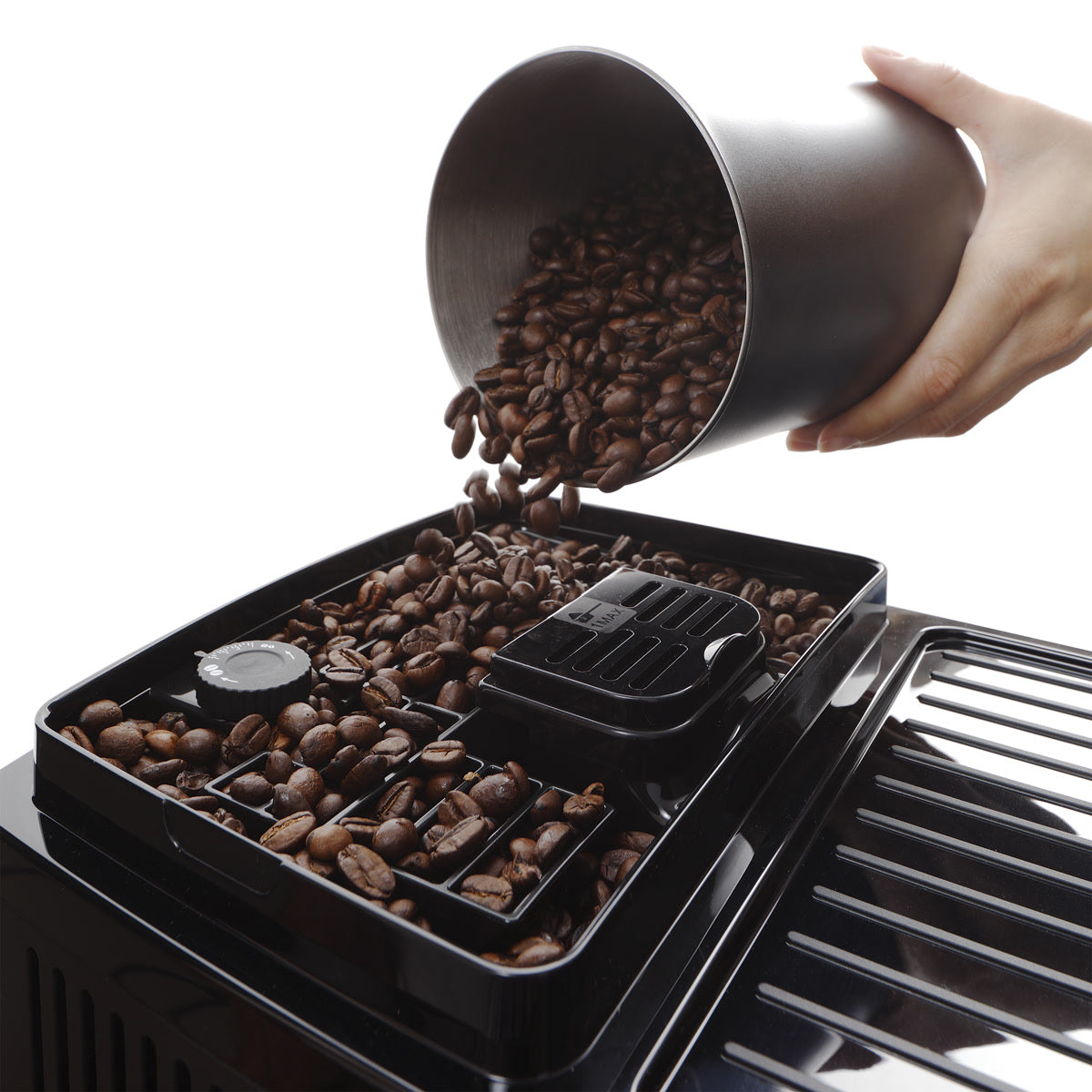 A hand pours coffee beans from a metal container into the bean hopper of a DeLonghi Magnifica Start Super Automatic Espresso Machine ECAM22022SB (Silver), preparing them for the integrated conical burr grinder.