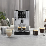 The DeLonghi Magnifica Start Super Automatic Espresso Machine ECAM22022SB (Silver) with a conical burr grinder brews two cups of coffee, surrounded by mugs featuring various coffee drinks and a metal milk frothing pitcher, on a light kitchen counter.