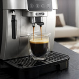 The De'Longhi Magnifica Start Super Automatic Espresso Machine ECAM22022SB in silver, featuring a conical burr grinder, pours coffee into a clear glass cup on its black drip tray, while a living room with a sofa appears blurred in the background.