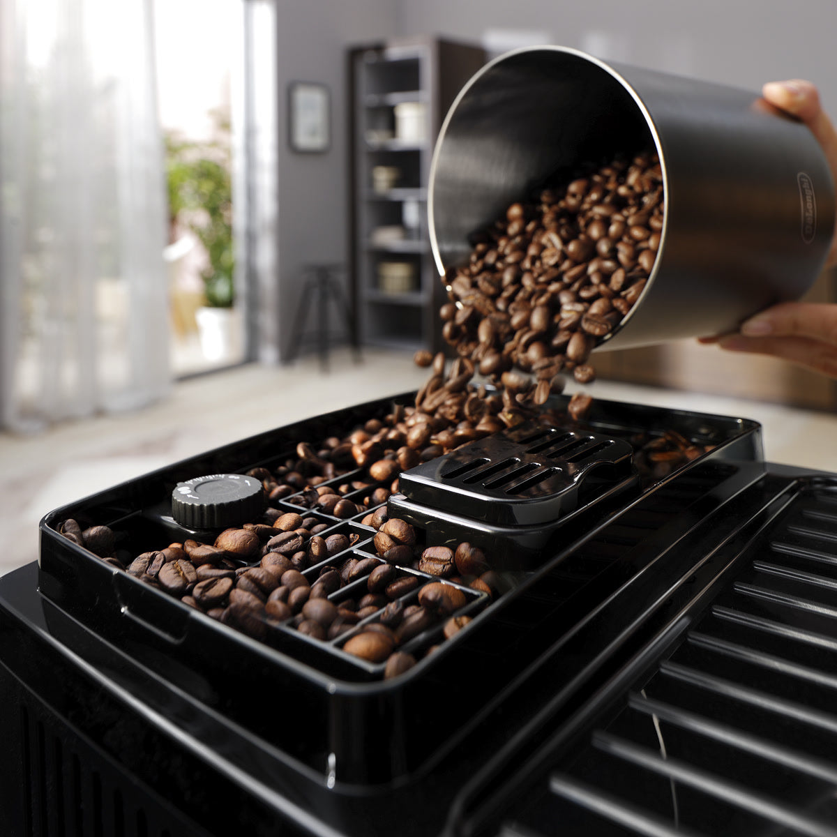 A hand pours coffee beans from a metal container into the hopper of the De'Longhi Magnifica Start Super Automatic Espresso Machine ECAM22022SB (Silver) by DeLonghi, which features a built-in conical burr grinder and is set in a modern, sunlit kitchen.
