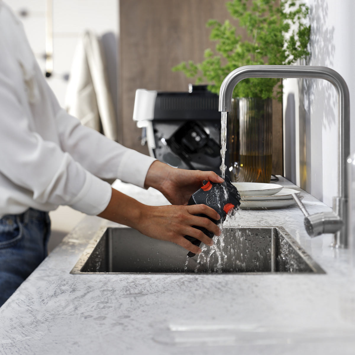 A person rinses a part of the De'Longhi Magnifica Start Super Automatic Espresso Machine ECAM22022SB (Silver) by DeLonghi under running water at a kitchen sink, with plates and a small plant visible in the background.