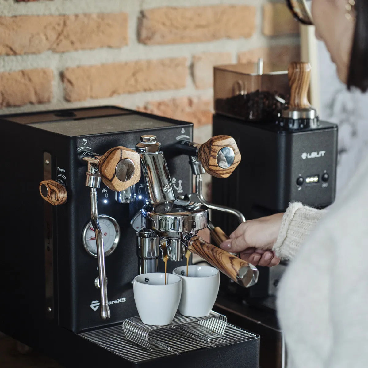 A person brews coffee into two white cups using the Lelit Mara X3 Semi-Automatic Heat-Exchange Espresso Machine PL62X3 (Black) by Lelit, with a coffee grinder and brick wall in the background.