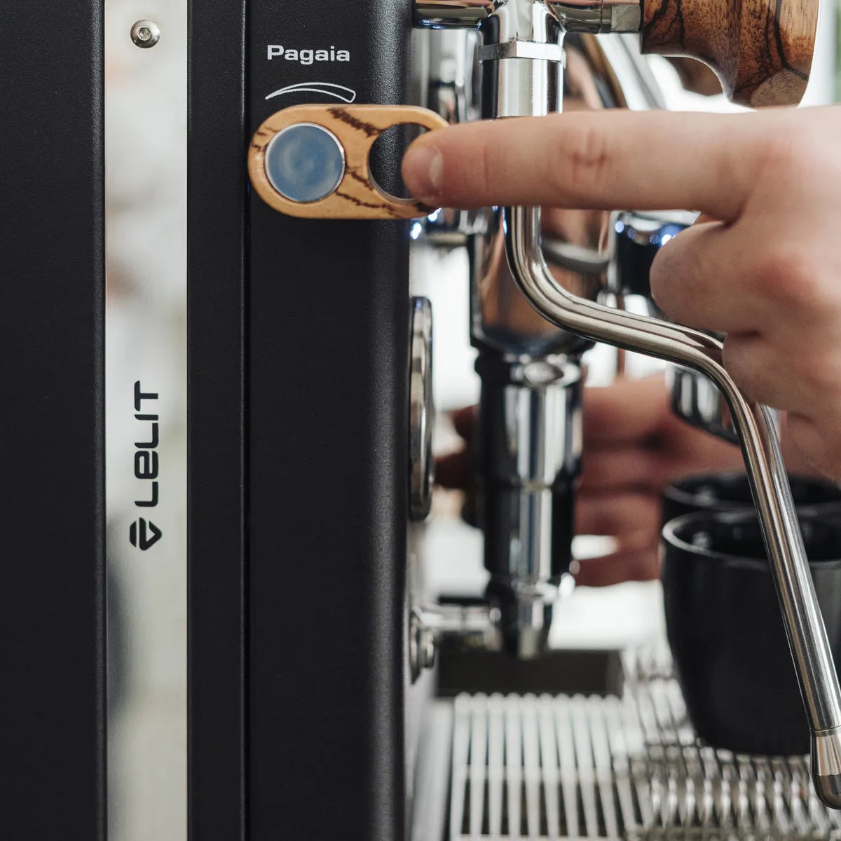 A hand presses the wooden "Pagaia" button on a Lelit Mara X3 Semi-Automatic Heat-Exchange Espresso Machine PL62X3 (Black), with its steam wand and black cups in the background.
