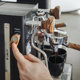 A close-up of a person turning a wooden knob on the Lelit Mara X3 Semi-Automatic Heat-Exchange Espresso Machine PL62X3 (Black) as espresso pours into a black cup below the portafilter.