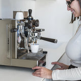 A woman in glasses and a light top smiles as she makes coffee with the Lelit Mara X3 Semi-Automatic Heat-Exchange Espresso Machine PL62X3 (Stainless Steel), watching espresso pour into a white cup on the drip tray of her modern kitchen.