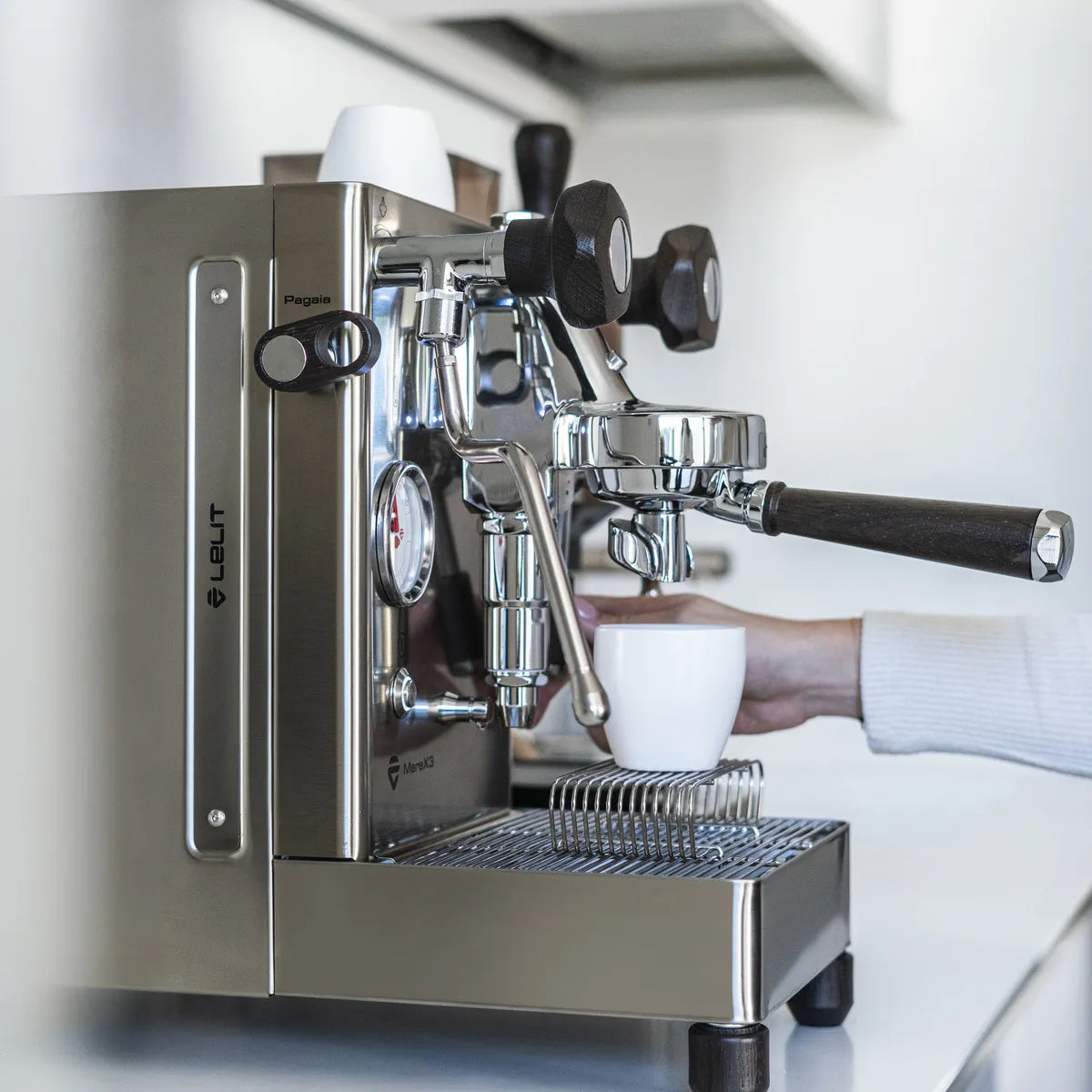 A person holds a white cup under the spout of the stainless steel Lelit Mara X3 Semi-Automatic Heat-Exchange Espresso Machine PL62X3, preparing to make coffee on a kitchen counter.