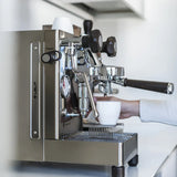 A person holds a white cup under the spout of the stainless steel Lelit Mara X3 Semi-Automatic Heat-Exchange Espresso Machine PL62X3, preparing to make coffee on a kitchen counter.