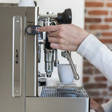 A hand presses a button on the Lelit Mara X3 Semi-Automatic Heat-Exchange Espresso Machine PL62X3 (Stainless Steel), preparing coffee in a white cup, with an exposed brick wall in the background.