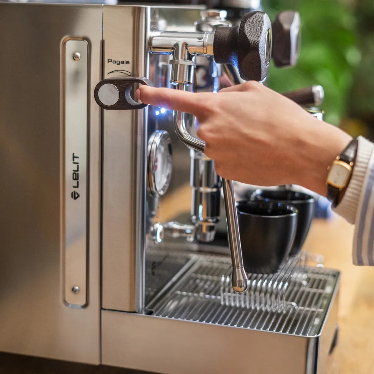 A hand wearing a watch presses a button on the stainless steel Lelit Mara X3 Semi-Automatic Heat-Exchange Espresso Machine PL62X3, with two black cups placed on the drip tray beneath the steam wand.