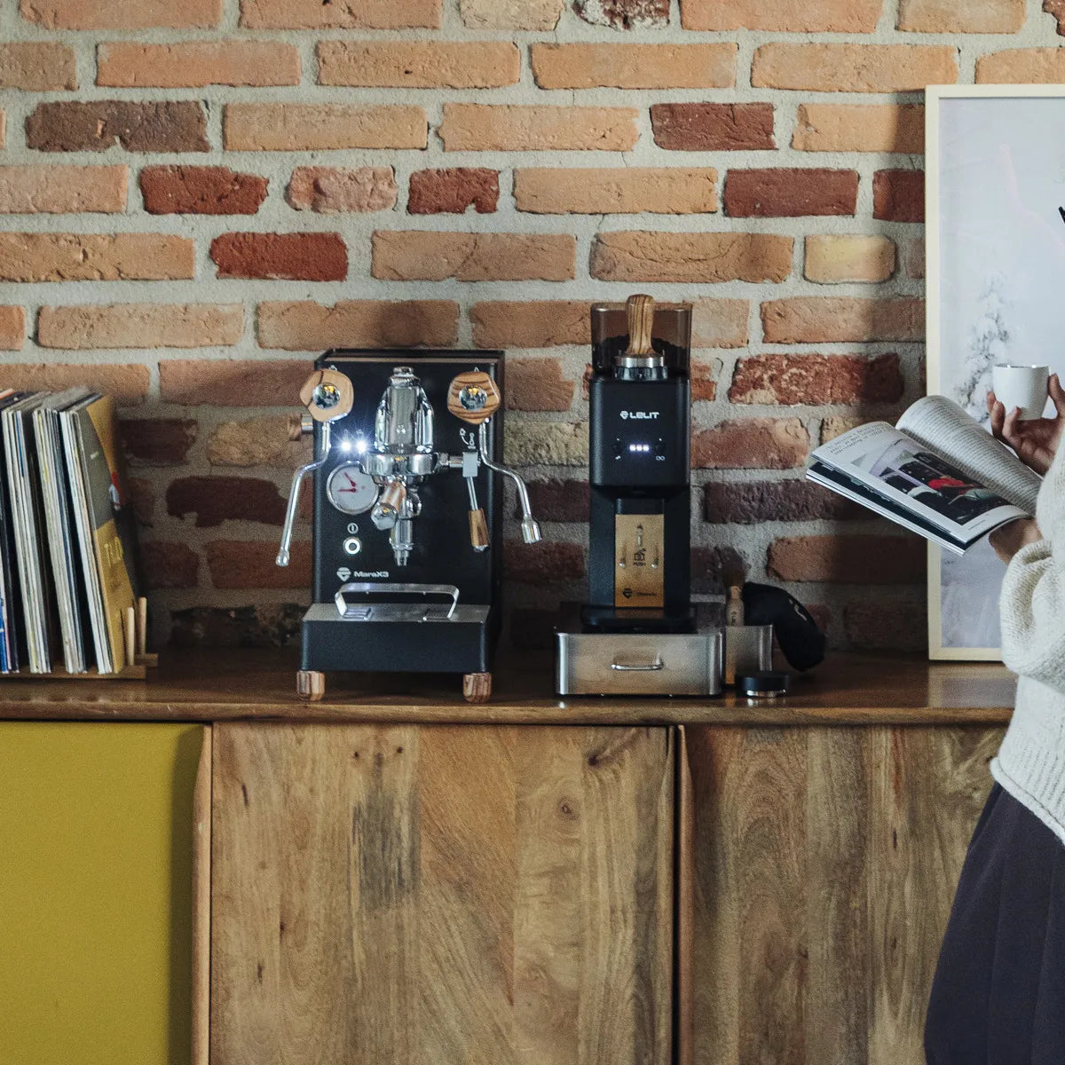 A person reads a book beside a wooden cabinet holding a Lelit Mara X3 Semi-Automatic Heat-Exchange Espresso Machine PL62X3 (Black) by Lelit, a coffee grinder, and vinyl records against a brick wall.
