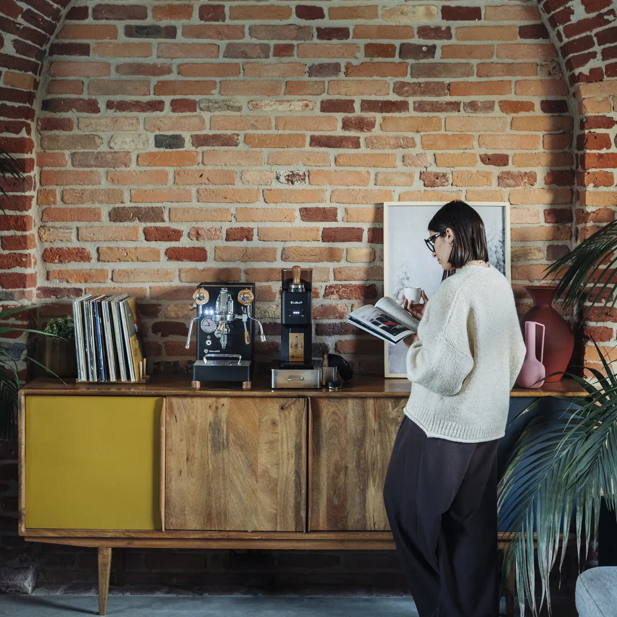 A person in a white sweater stands by a wooden sideboard with the Lelit Mara X3 Semi-Automatic Heat-Exchange Espresso Machine PL62X3 (Black), records, and decor against an exposed brick wall, reading a book in this cozy, plant-filled room.