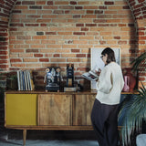 A person in a white sweater stands by a wooden sideboard with the Lelit Mara X3 Semi-Automatic Heat-Exchange Espresso Machine PL62X3 (Black), records, and decor against an exposed brick wall, reading a book in this cozy, plant-filled room.