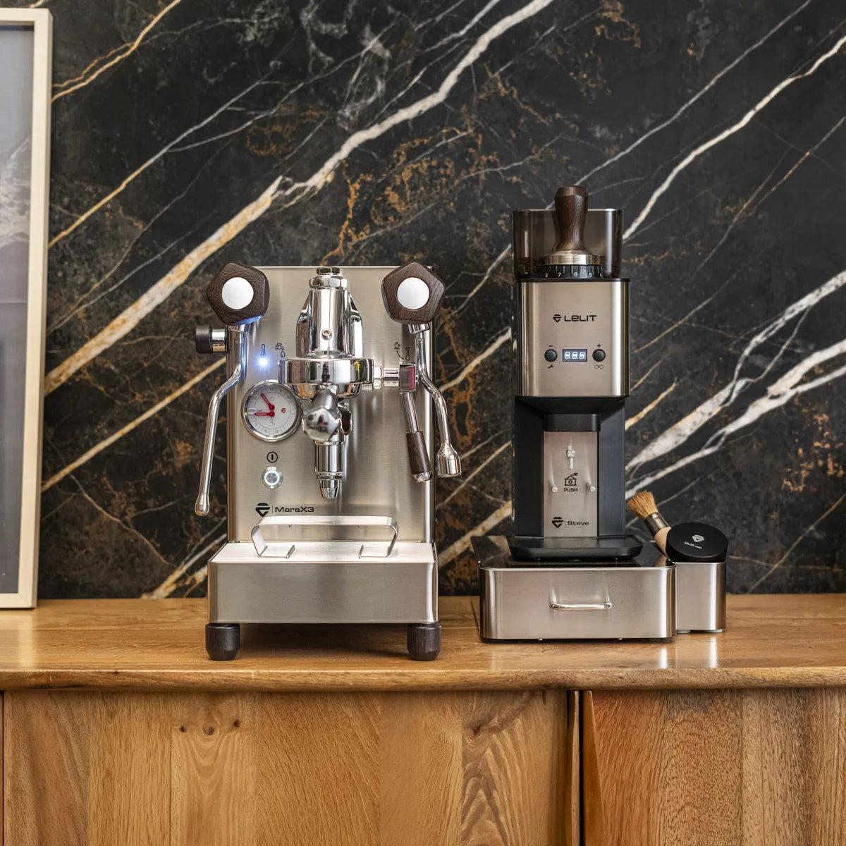 A Lelit Mara X3 Semi-Automatic Heat-Exchange Espresso Machine PL62X3 (Stainless Steel) by Lelit and a coffee grinder are placed side by side on a wooden counter before a black marble wall with white and gold veins.
