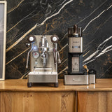 A Lelit Mara X3 Semi-Automatic Heat-Exchange Espresso Machine PL62X3 (Stainless Steel) by Lelit and a coffee grinder are placed side by side on a wooden counter before a black marble wall with white and gold veins.