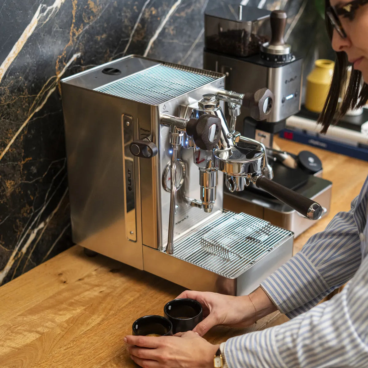 Someone prepares espresso with a Lelit Mara X3 Semi-Automatic Heat-Exchange Espresso Machine PL62X3 (Stainless Steel) by Lelit on a wooden countertop, placing two small black cups under the portafilter with coffee accessories in the background.