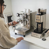 A woman makes espresso using the Lelit Mara X3 Semi-Automatic Heat-Exchange Espresso Machine PL62X3 (Stainless Steel) beside a grinder on a white countertop, with cups, a tamper, and electronic flow control also visible.