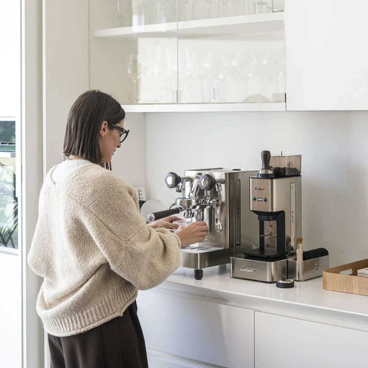 A person with shoulder-length dark hair and glasses uses the Lelit Mara X3 Semi-Automatic Heat-Exchange Espresso Machine PL62X3 (Stainless Steel) in a modern white kitchen with glassware on shelves above the counter.
